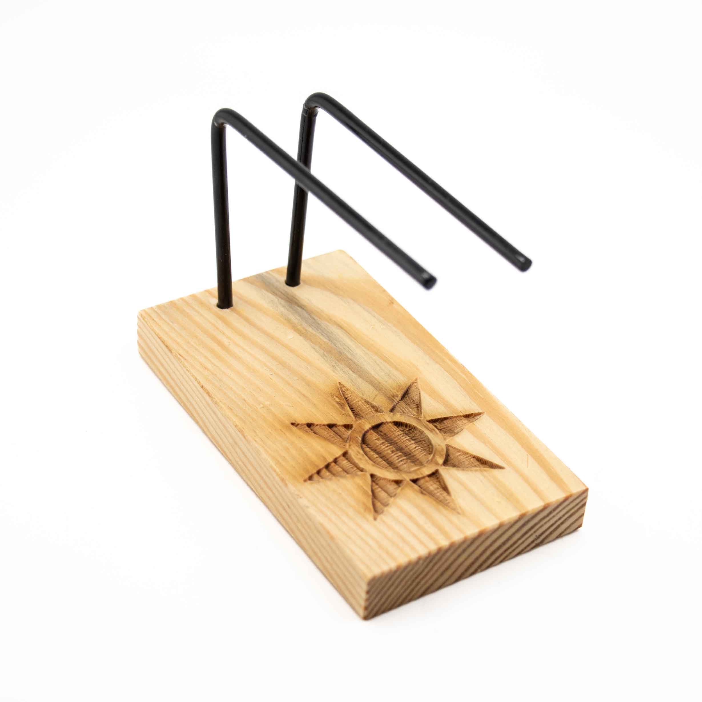 A wooden smudge stick burner featuring a light-grained rectangular base with a carved sun design and two black metal support rods.