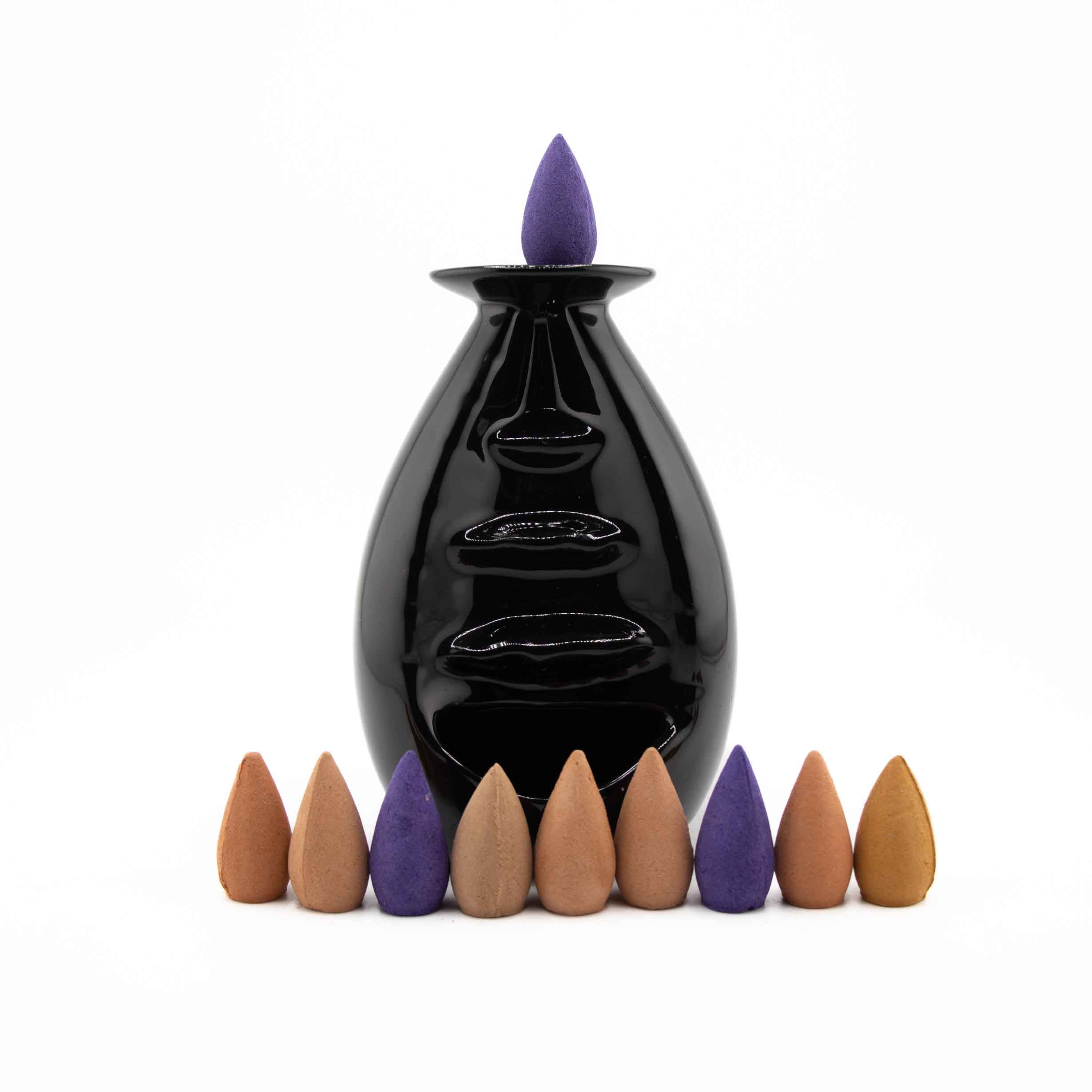 A glossy black ceramic backflow incense burner shaped like a face, featuring a purple incense cone on top and a row of colorful cones at the base.